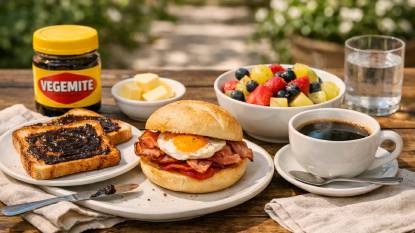 A classic Australian breakfast table featuring Vegemite on toast, a bacon and egg roll, a bowl of fresh fruit, and a cup of strong coffee or tea, set on a sunny patio
