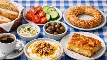 A Greek breakfast table with a small cup of strong Greek coffee, thick strained yogurt drizzled with honey and walnuts, fresh bread, olives, tomatoes, a slice of tiropita, and a koulouri sesame bread ring on a blue-and-white tablecloth
