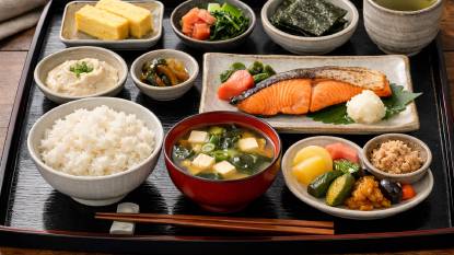 A traditional Japanese breakfast set with a bowl of steamed rice, miso soup with tofu and seaweed, grilled salmon fillet, pickled vegetables, a small tamagoyaki omelette, and green tea arranged on a lacquer tray