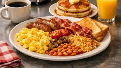 An American breakfast spread with fluffy pancakes stacked high with butter and maple syrup, crispy bacon strips, scrambled eggs, a glazed doughnut, a toasted bagel with cream cheese, a glass of orange juice, and a mug of coffee on a diner counter