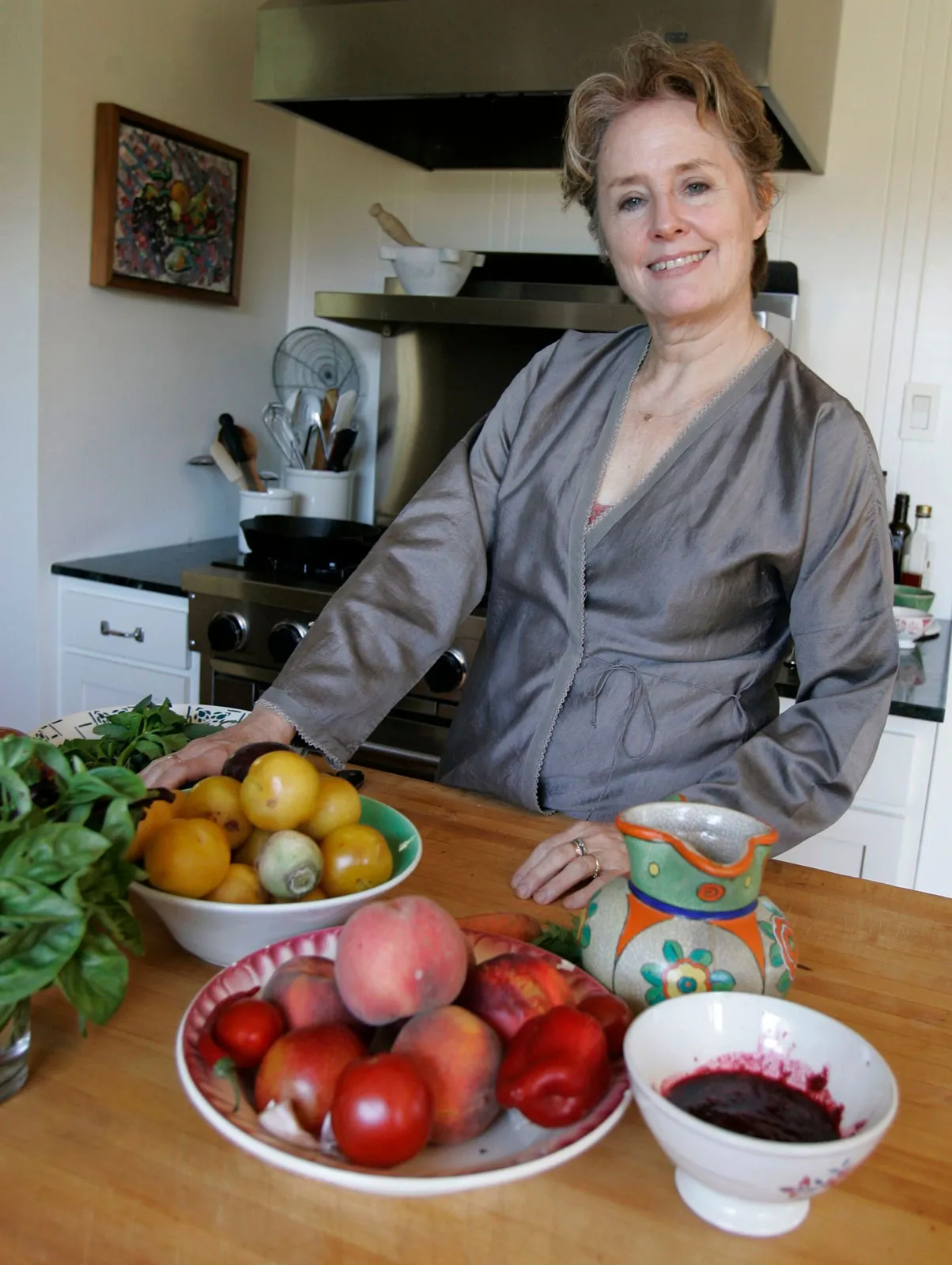 Chef Alice Waters standing in a bright kitchen, surrounded by fresh seasonal produce, looking toward the camera with a calm, thoughtful expression
