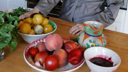 Chef Alice Waters standing in a bright kitchen, surrounded by fresh seasonal produce, looking toward the camera with a calm, thoughtful expression