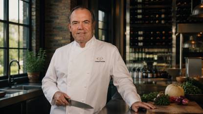 Chef Charlie Palmer. An AI‑generated artistic impression of American chef Charlie Palmer, depicted in a 16:9 editorial‑style composition. He has short silver‑grey hair and stands in a modern kitchen with exposed brick and stainless steel counters. He wears a white chef’s coat, holding a chef’s knife near a cutting board with fresh produce. Soft window light creates warm shadows and highlights the metallic and organic textures. The image is photorealistic and cinematic, with amber and slate tones. This is a stylized imaginative rendering, not an actual photograph.