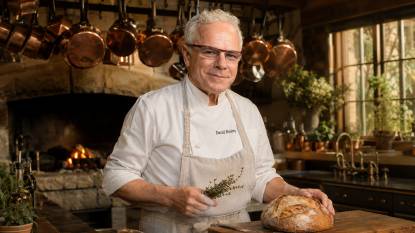 Chef David Bouley. An AI‑generated artistic impression of American chef David Bouley, depicted in a 16:9 editorial‑style composition. He has salt‑and‑pepper hair, a mustache, and a focused expression. He stands in a rustic French‑inspired kitchen with wooden beams, a stone hearth, and hanging copper pots. He wears a white chef’s coat and an apron, holding a loaf of sourdough bread and fresh thyme. Warm afternoon light creates soft golden shadows, highlighting the copper and wood textures. The image is photorealistic and cinematic, with amber and olive tones — this is a stylized imaginative rendering, not an actual photograph.