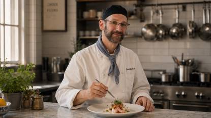 Chef David Waltuck. An AI‑generated artistic impression of American chef David Waltuck, depicted in a 16:9 editorial‑style composition. He has short graying hair and a gentle smile, standing in a clean, elegant kitchen with white subway tile walls and a marble counter. He wears a white chef’s coat with a blue‑gray neckerchief, holding a plate of delicately plated seafood and a pair of tweezers. Soft, diffused natural light creates a serene, airy mood with muted cream and slate‑blue tones. This is a stylized imaginative rendering, not an actual photograph.