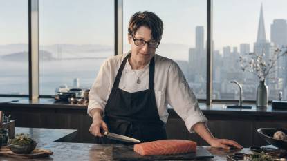 Chef Elka Gilmore. An AI‑generated artistic impression of American chef Elka Gilmore, depicted in a 16:9 editorial‑style composition. She has a short pixie haircut and a confident expression, standing in a bright San Francisco kitchen with large windows overlooking the city. She wears a white chef’s coat and a black apron, holding a Japanese knife beside a piece of raw fish on a slate board. Soft, foggy morning light creates cool blue‑gray tones with warm salmon highlights. This is a stylized imaginative rendering, not an actual photograph.