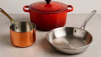 Copper saucepan, red enameled cast iron Dutch oven, and stainless steel frying pan arranged on a rustic kitchen counter with soft natural lighting.
