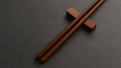 A pair of polished wooden chopsticks resting on a ceramic bowl atop a textured linen surface, softly lit to evoke the cultural history and etiquette associated with chopstick use.
