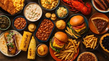 Overhead image of a buffet featuring iconic American dishes arranged on rustic wooden surfaces. Visible items include fried catfish, macaroni and cheese, barbecue beans, lobster, shrimp and grits, brisket, clam chowder, cheeseburgers with fries, fish tacos, collard greens, cornbread muffins, baked beans, blueberry pie, and pecan pie. The composition highlights regional diversity and comfort food classics in a warm, editorial setting.