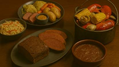 four traditional New England dishes arranged on a dark wooden table. On the left, a sliced loaf of steamed brown bread with a cracked top and dense, molasses-rich interior sits on a dark gray ceramic plate. Next to it, Indian pudding with a deep brown color and slightly cracked surface is served in a reddish-brown earthenware bowl. Centered in the image, a New England boiled dinner is plated on a round teal dish, featuring thick slices of corned beef, boiled cabbage, potatoes, and carrots, with a small bowl of mustard above the meat. On the right, a seafood clambake is presented in a large metal pot filled with bright red lobsters, open clams, and yellow corn on the cob. The lighting is warm and soft, emphasizing the rustic textures and earthy tones of the dishes