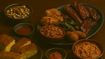 American soul food arranged on a dark wooden table. At the center, a blue ceramic plate features glazed barbecue ribs with a caramelized crust, golden fried chicken with a crisp coating, and a mound of slow-cooked collard greens. In the foreground, rectangular slices of cornbread with a golden crust and crumbly yellow interior are placed on a round dark platter. To the right, reddish-brown ceramic bowls contain red beans and rice, golden hush puppies, and creamy sweet mashed potatoes. On the left, a rustic bowl holds black-eyed peas topped with simmered pork, and a smaller bowl of Tabasco sauce sits nearby. The dishes are illuminated with warm, directional lighting that highlights their textures and rich colors, evoking warmth, tradition, and communal celebration.